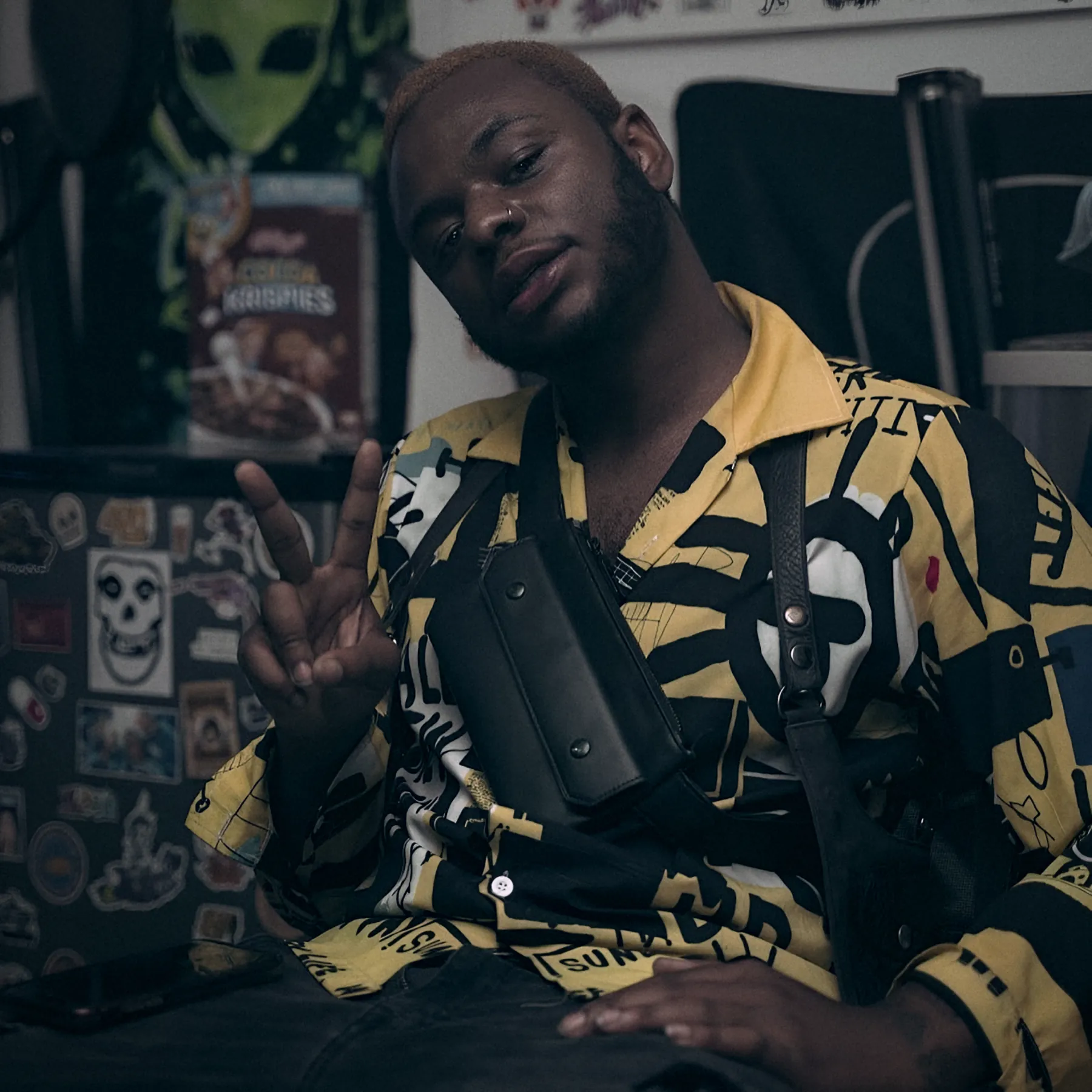 Bobby Brown Jr. in a yellow-and-black shirt, seated and throwing a peace sign beside an alien poster