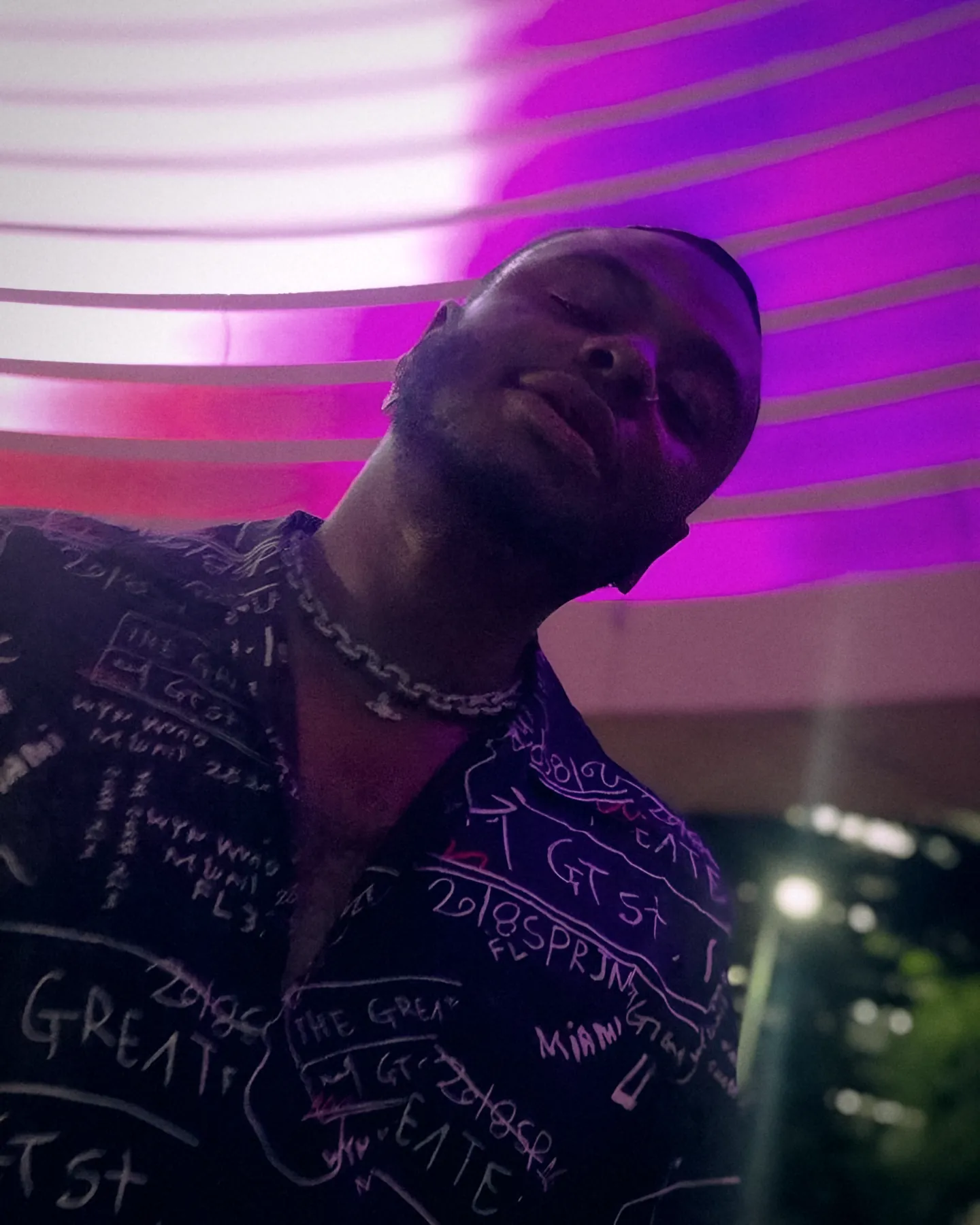 Bobby Brown Jr. tilting his head under pink and purple neon stripes, wearing a black 'The Greatest' print shirt