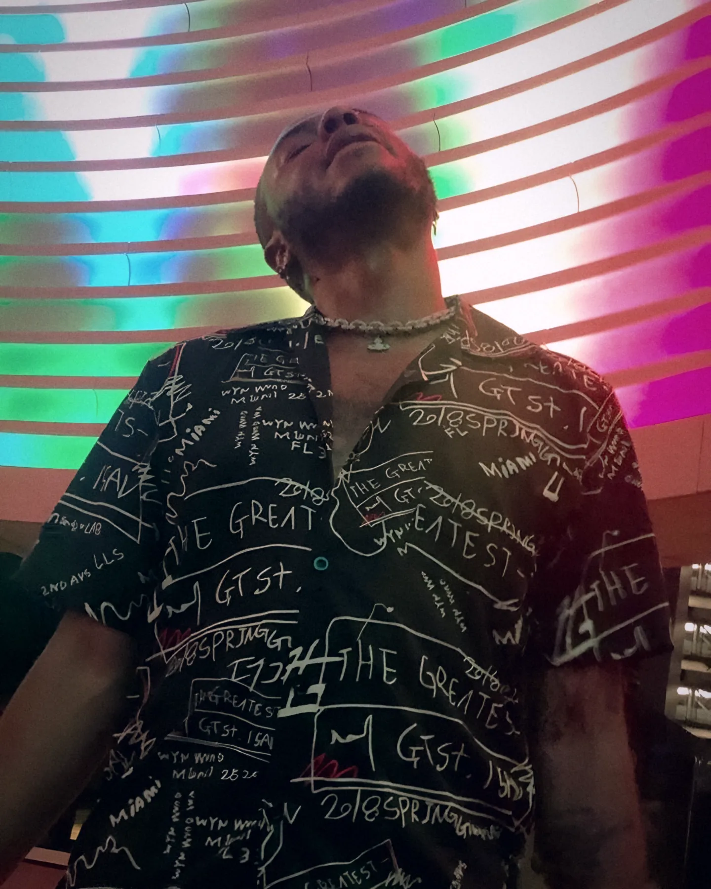 Bobby Brown Jr. looking up beneath a striped neon ceiling in a black 'The Greatest' print shirt