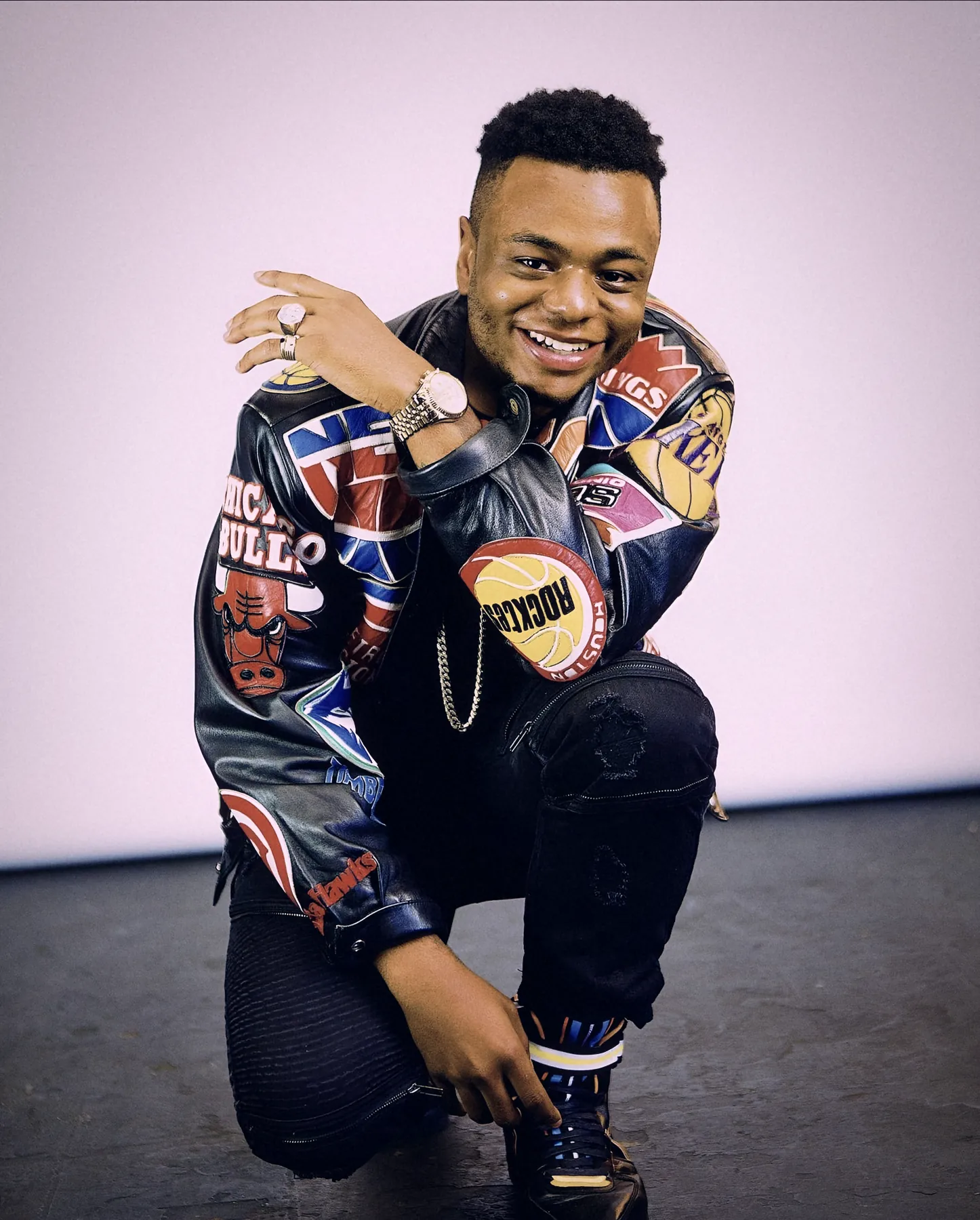 Bobby Brown Jr. kneeling and smiling in a patch-covered leather jacket against a white studio wall